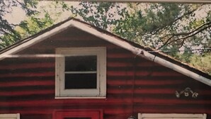 A red cabin with a heart-shaped decoration on the door, a small porch with a grill, and a vase with flowers. - Peaceful Vintage 1920's Lakefront Red Log Cabin (Fifty Lakes)