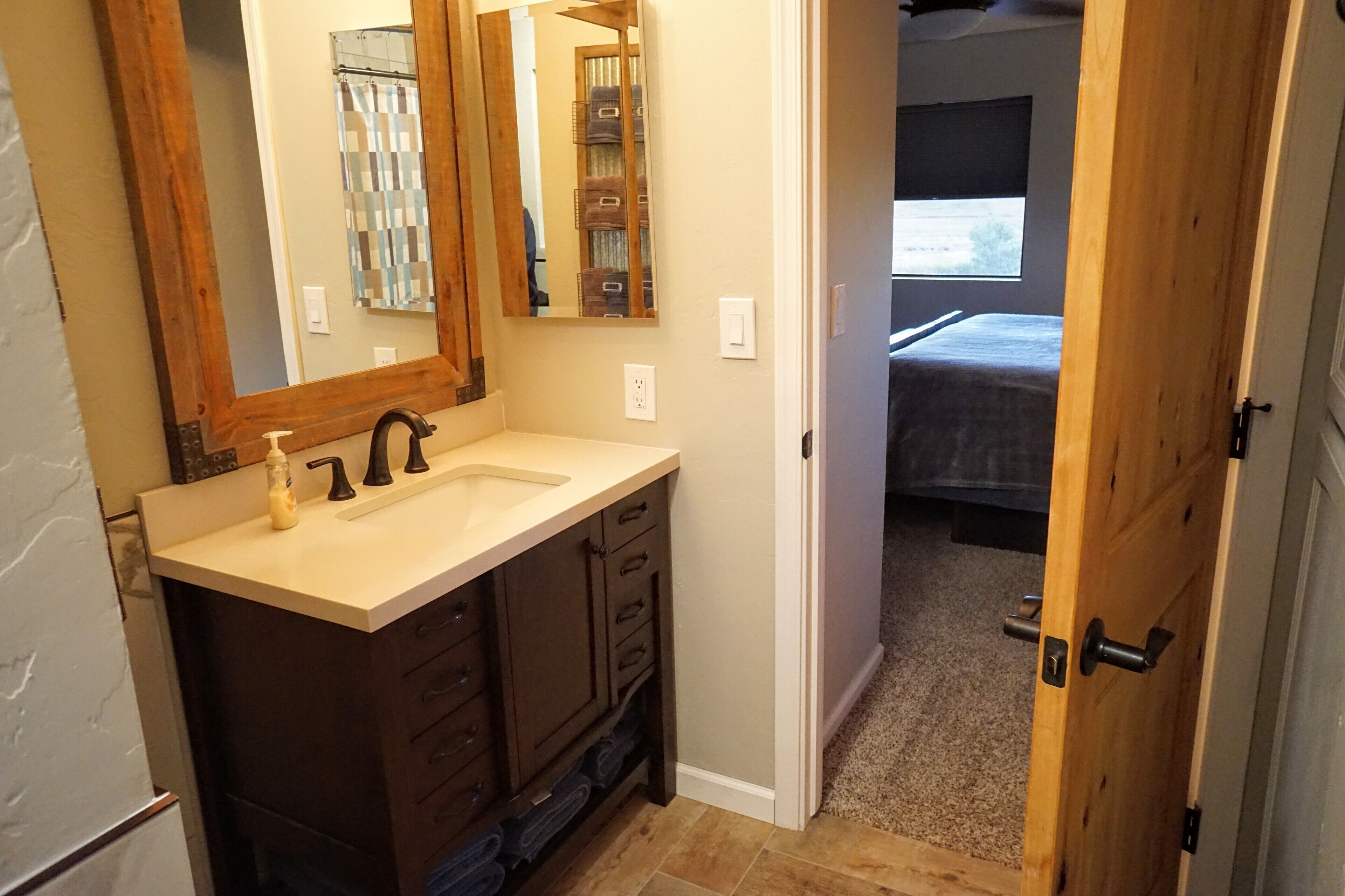 A bathroom with a wooden vanity, a large mirror, and a view into a bedroom through an open door.