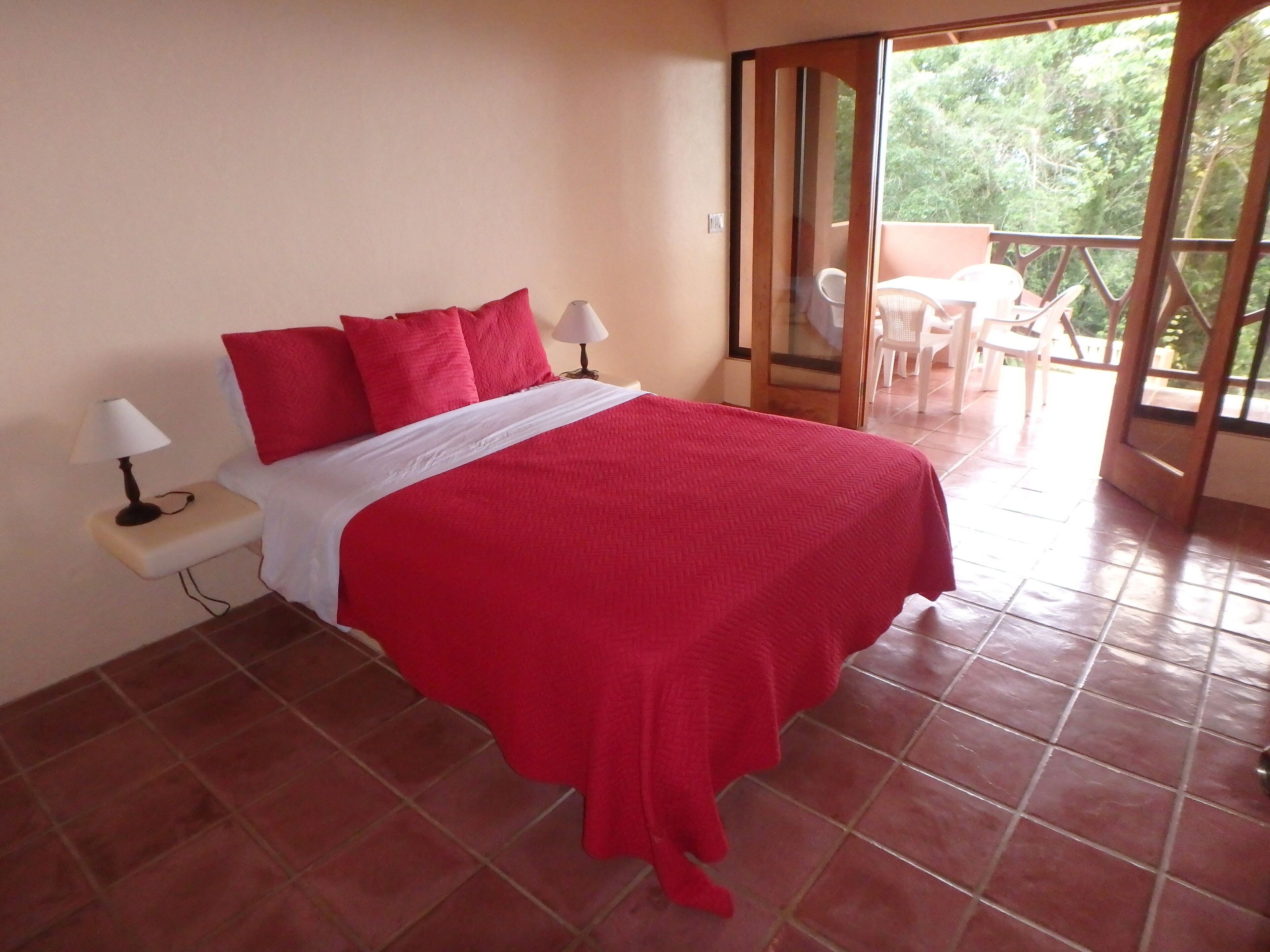 Een slaapkamer met een rood beddekleed, twee bedlampen en uitzicht op een balkon met stoelen.