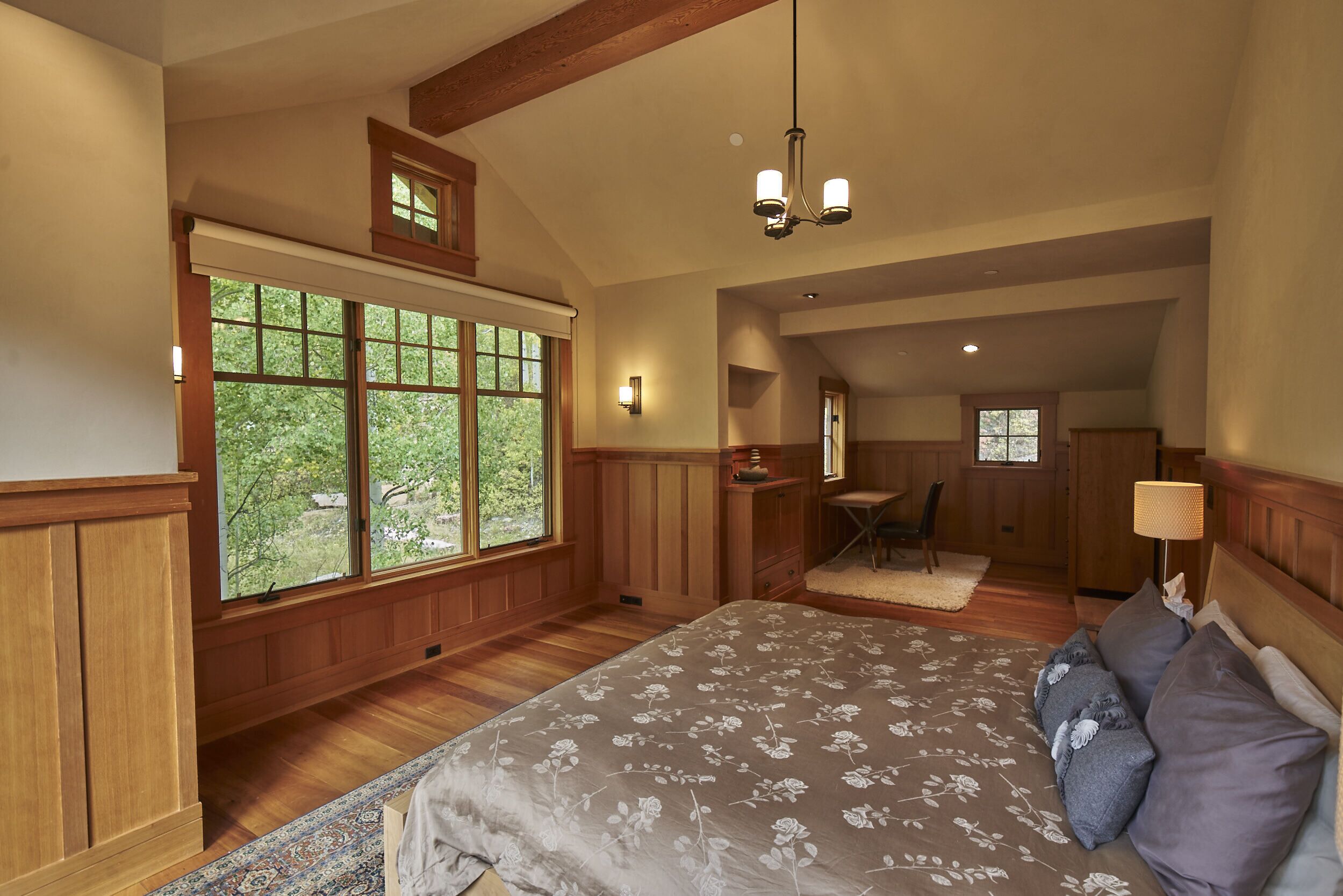 A bedroom with a large bed, wooden paneled walls, a window with a view of trees, and a small desk area.
