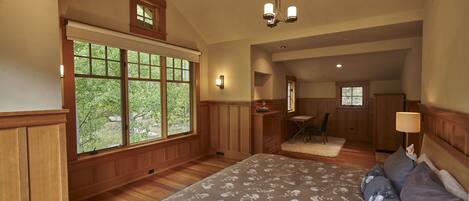 A bedroom with a large bed, wooden paneled walls, a window with a view of trees, and a small desk area.