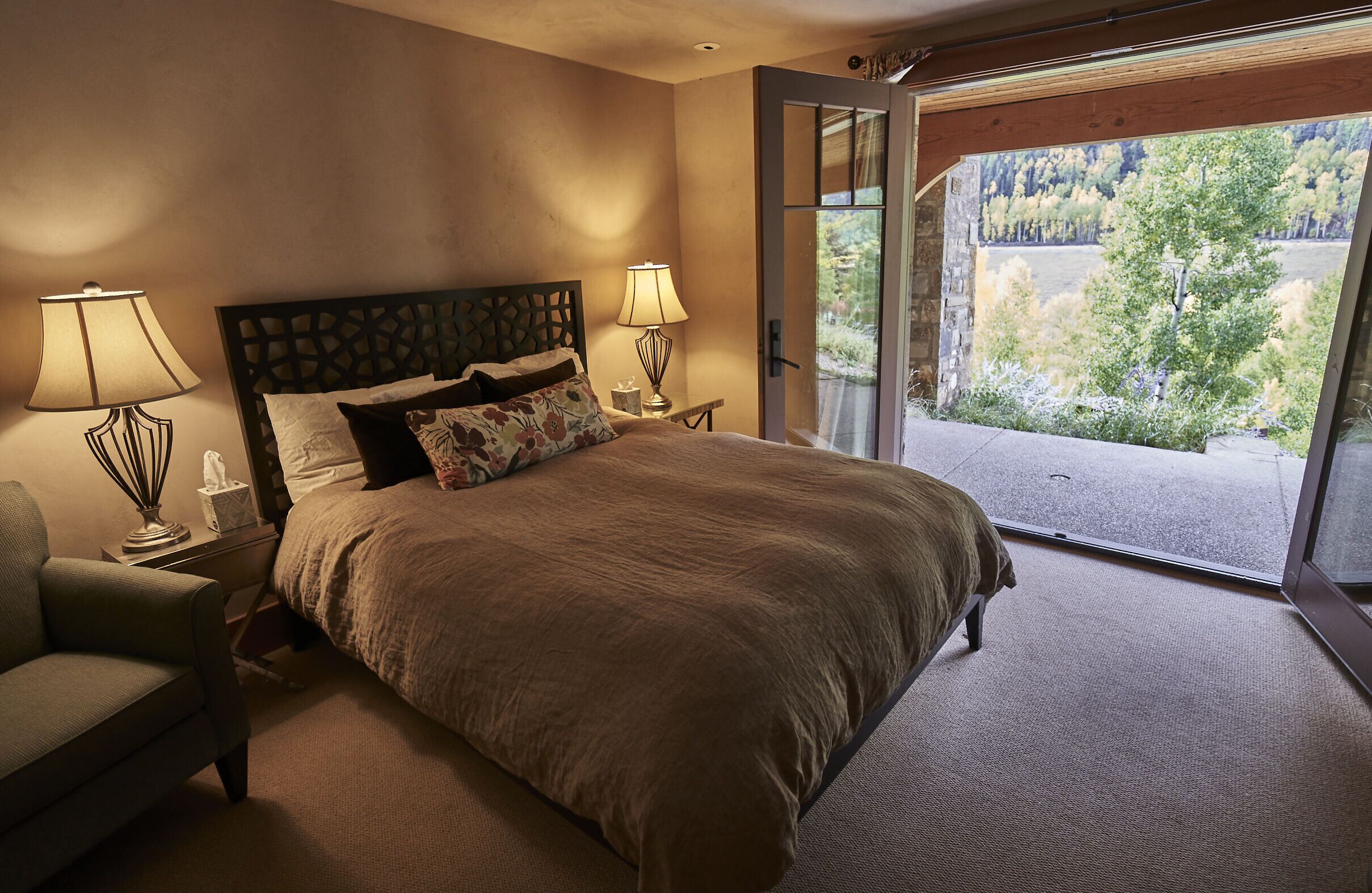 Camera da letto con un letto grande, due lampade sui comodini, una sedia e vista su una foresta attraverso una porta in vetro.