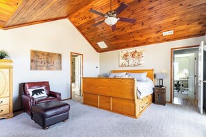 Une chambre avec un plafond en bois, un lit, une chaise avec un coussin en forme d’ours et un tableau accroché au mur.