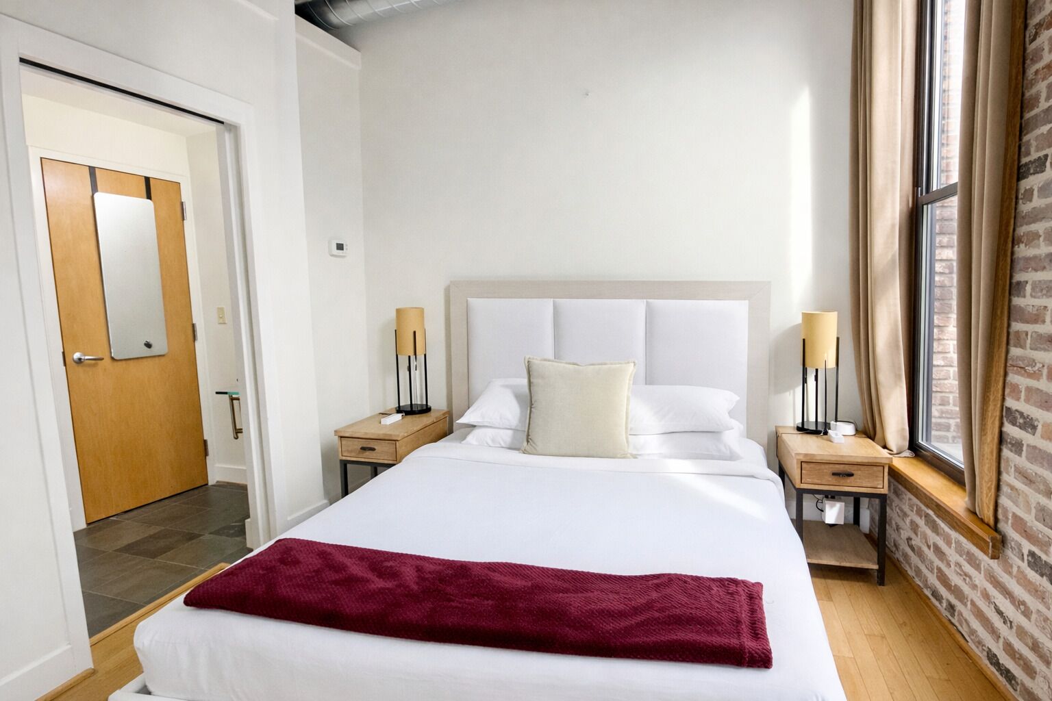 A hotel room with a large bed, two bedside tables, a wooden door, a brick wall, and a window with curtains.