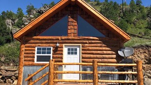 A log cabin with a wooden deck, two windows, a door, and a satellite dish. - 8 miles to Mount Rushmore - Sleeps 2 to 6 (Rapid City)