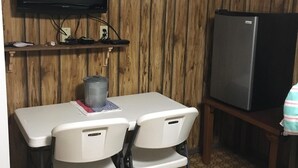Ett litet rum med en väggpanel av trä, en platt-TV som är monterad ovanför ett litet bord med två stolar, ett kylskåp och en papperskorg.
