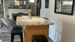 A kitchen with a granite countertop, two black stools, a hanging light fixture, and a brick fireplace. - Archie's Lake Daze Cottage - No Pets (Manning)
