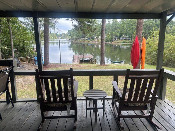 A wooden deck with two rocking chairs and a small table, overlooking a calm lake with dock and paddle boats.