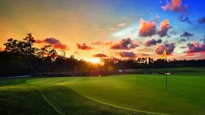 Un campo de golf con una bandera al final del callejón, rodeado de árboles y un cielo al atardecer.