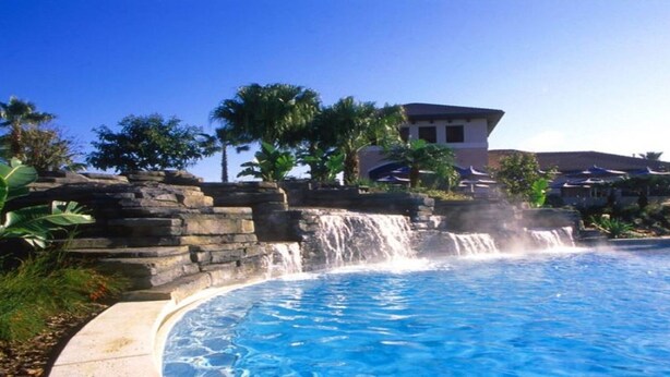 Una piscina con cascada, rodeada de muros de piedra y plantas tropicales.