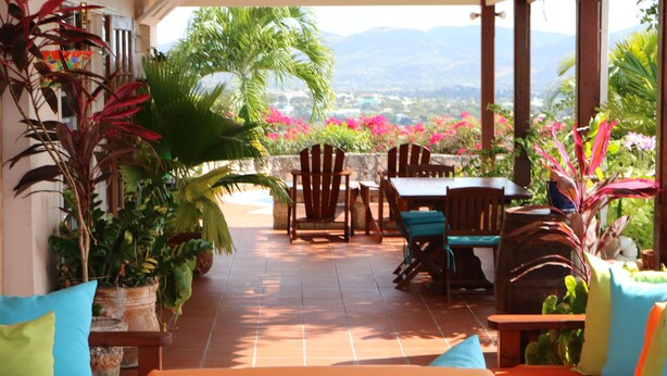 A covered patio with wooden furniture, potted plants, and a view of the landscape.