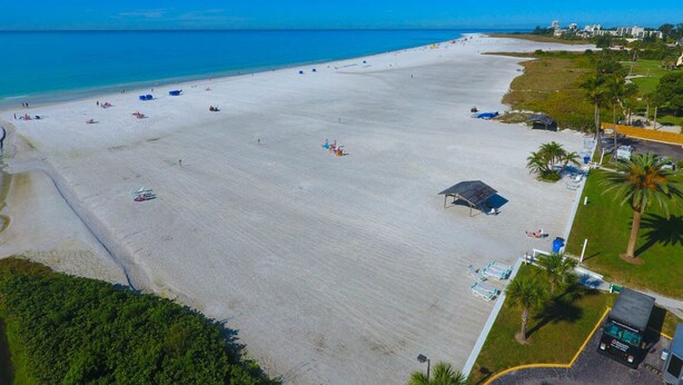Vista aérea de una playa con una extensa zona arenosa, algunos sombrillas y algunas personas. Se observan edificios al fondo y un estacionamiento con algunos automóviles.