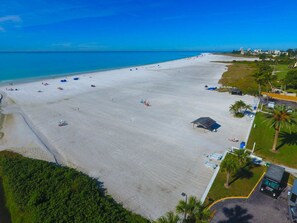 An aerial view of a beach with a large sandy area, a few umbrellas, and a few people. There are buildings in the background and a parking area with a few cars. - Siesta Key Gulf Side - #84 - Private Tranquil Condo - Jamaica Royale - Remodeled (Siesta Key)