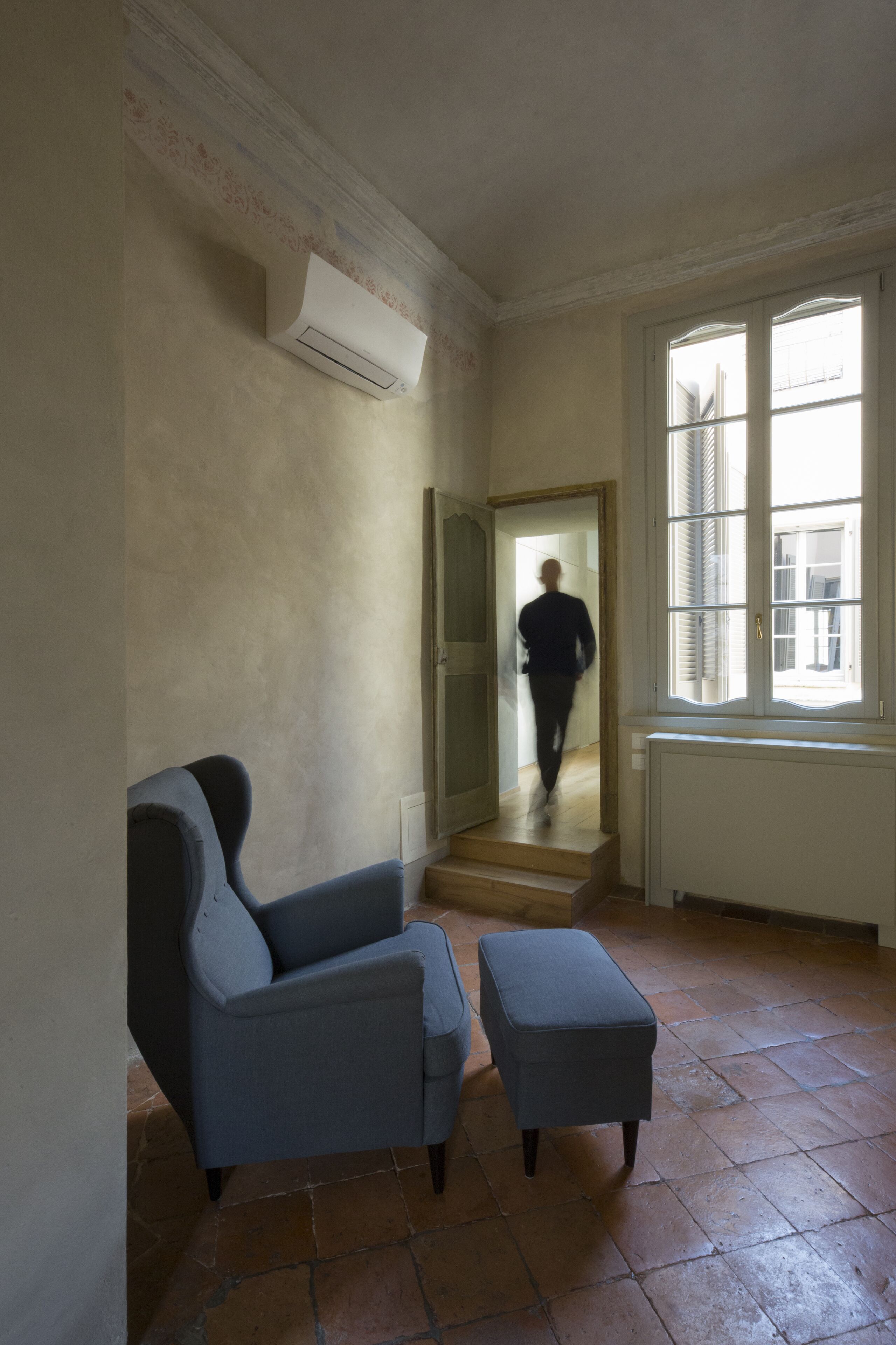 Une pièce comprenant une chaise, un pouf, une fenêtre et une porte donnant sur une autre pièce.
