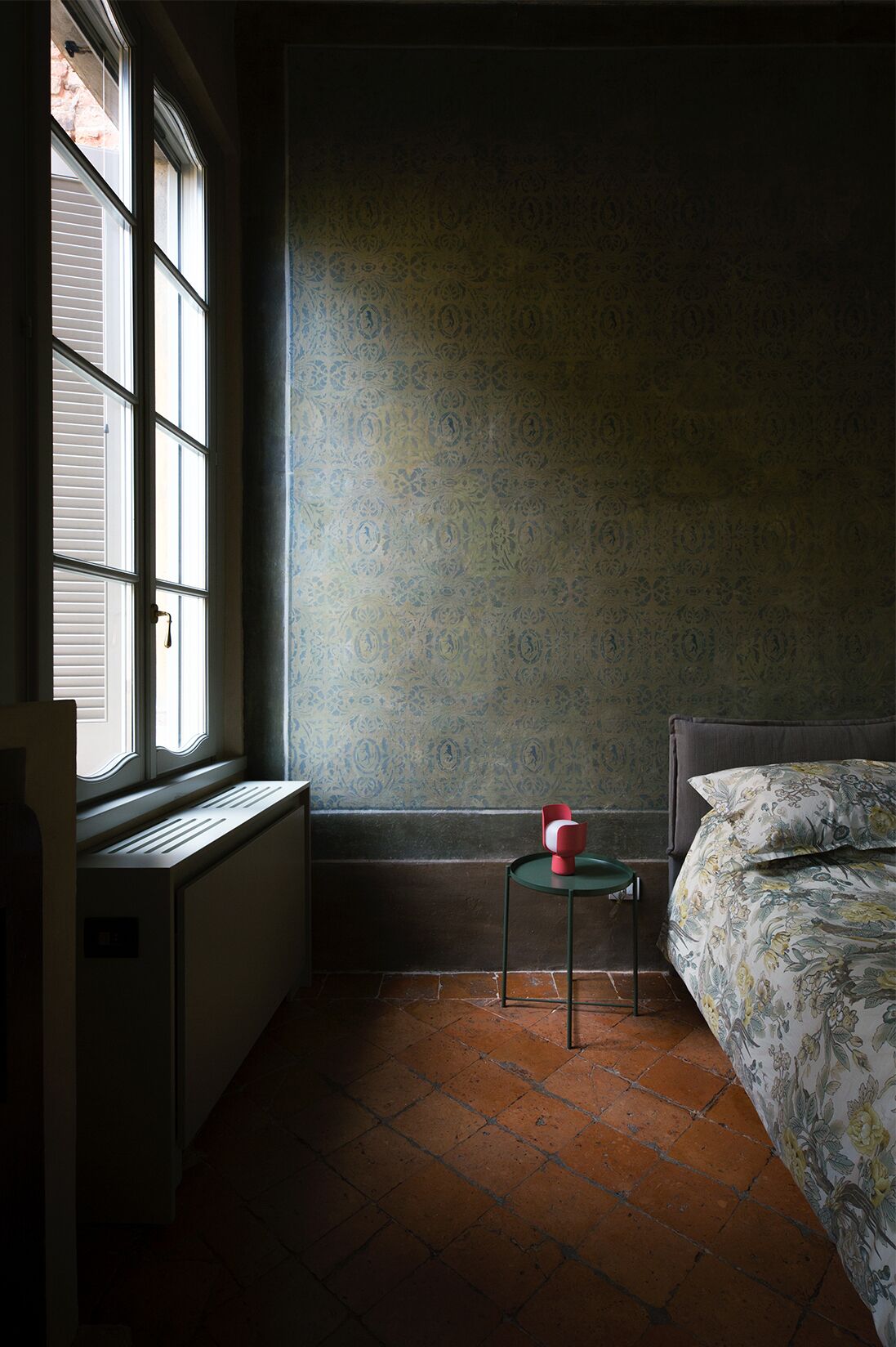 Una habitación con un radiador, una ventana, una pequeña mesa con un objeto rojo y una cama con ropa de cama estampada.