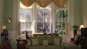 A cozy living room with a green sofa, a small table, and a chair. There are large windows with curtains, and a view of buildings outside. - Elegant Back Bay Brownstone-Comm Av 1+ Bed - 1 Week Min, beyond 2 wks considered (Boston)