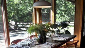 Una mesa de comedor dispuesta con platos, vasos y un jarrón de flores, rodeada de sillas rojas en una habitación con ventanas amplias.