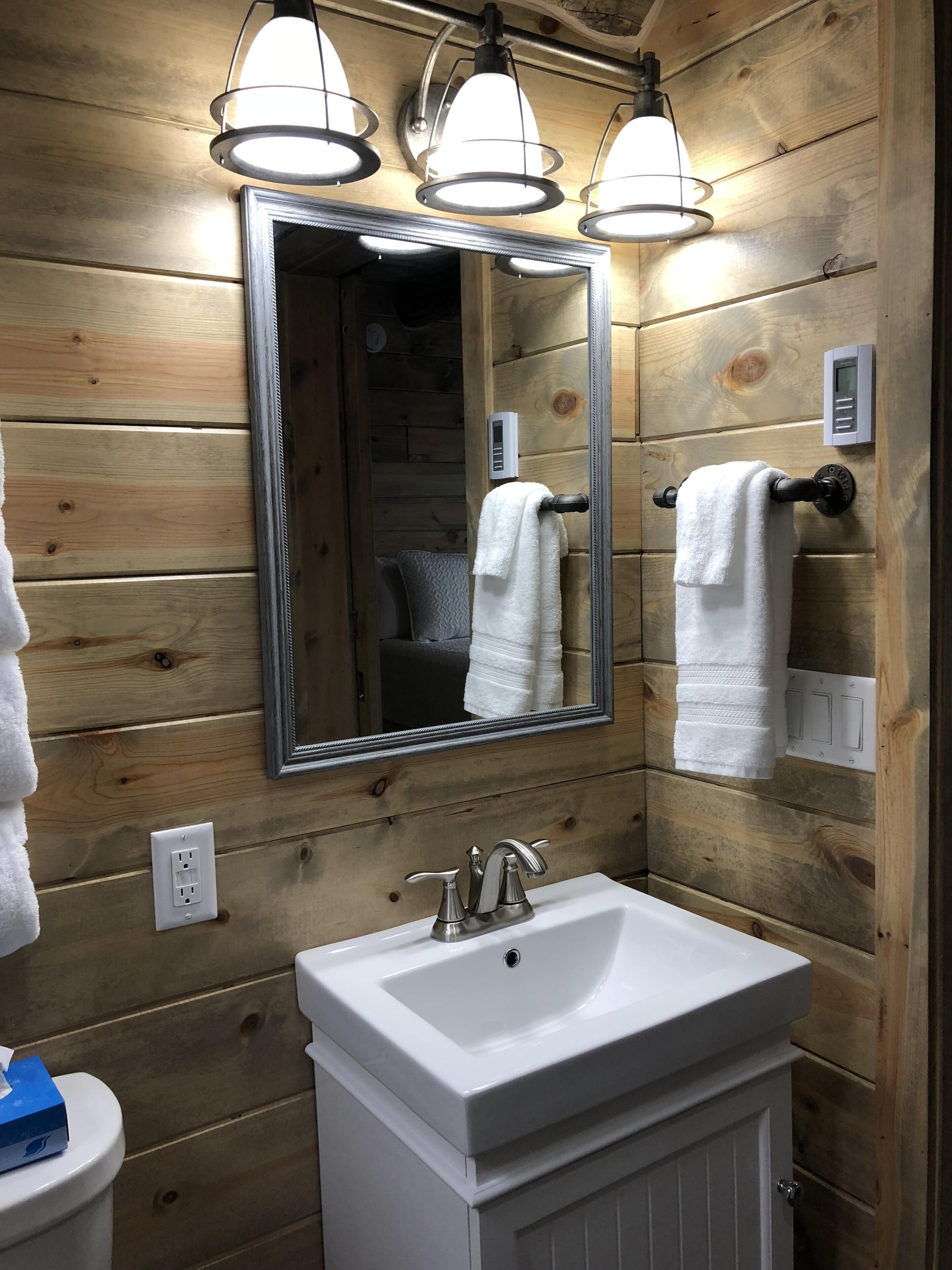A bathroom with wooden paneling, a large mirror, three pendant lights, a white sink, and two towel racks.