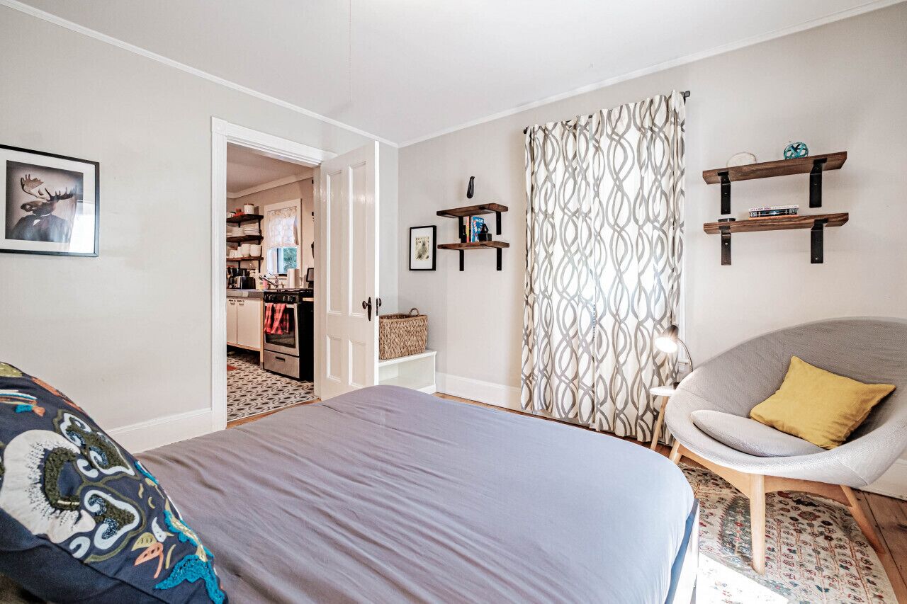 A bedroom with a bed, a chair, a patterned curtain, and shelves with books and decorative items.