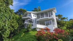 A modern two-story house with large windows and a balcony, surrounded by lush greenery and flowering plants. - Private Eco-Villa, Prime Central Location, Ocean Views, Wildlife, and Nightlife (Quepos)