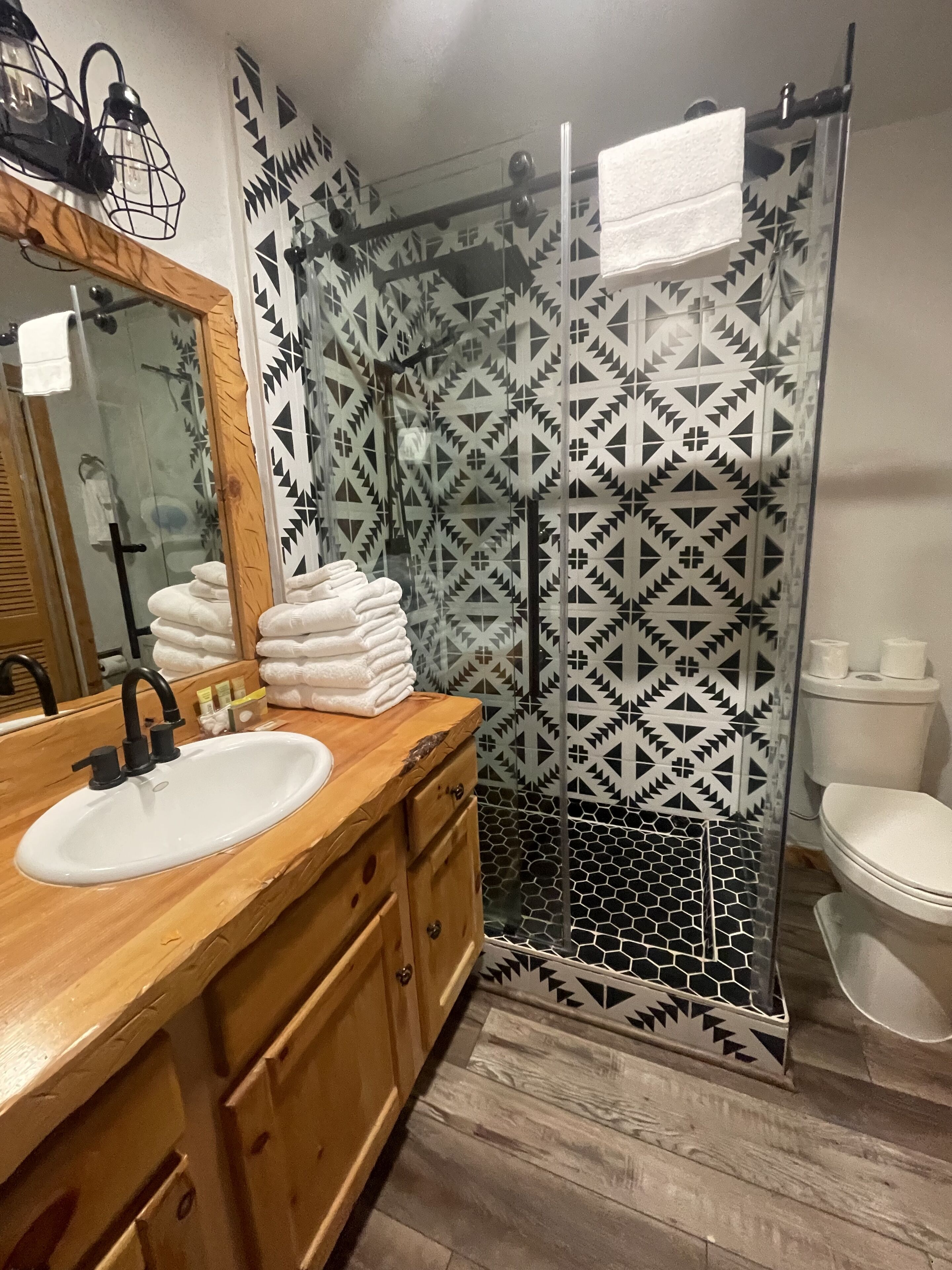 A bathroom with a wooden vanity, a sink, a mirror, a shower with a geometric tile pattern, and a toilet.