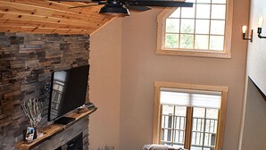 Ein geräumiges Wohnzimmer mit Kamin aus Stein, Ledersofas, Deckenventilator und großen Fenstern für natürliches Licht.