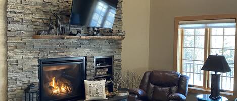 A living room with a stone fireplace, leather furniture, a flat-screen TV mounted on the wall, and a ceiling fan.