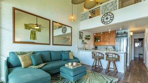 Uma sala de estar com sofá em forma de L, um banquinho e um espelho grande. Há uma área de cozinha com armários de madeira e um banco alto.