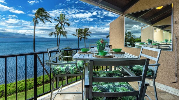 Varanda com mesa posta para refeições, com vista para um corpo d'água, palmeiras e montanhas ao fundo.