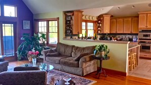 A living room with a sofa, a coffee table, and a small side table with a plant. There is a kitchen area with wooden cabinets and a countertop. - Newly Built 3 Story Home with Beach Parking Pass (Montauk)