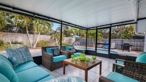 En overdækket terrasse med fletmøbler, et træbord og udsigt til en pool i baghaven.