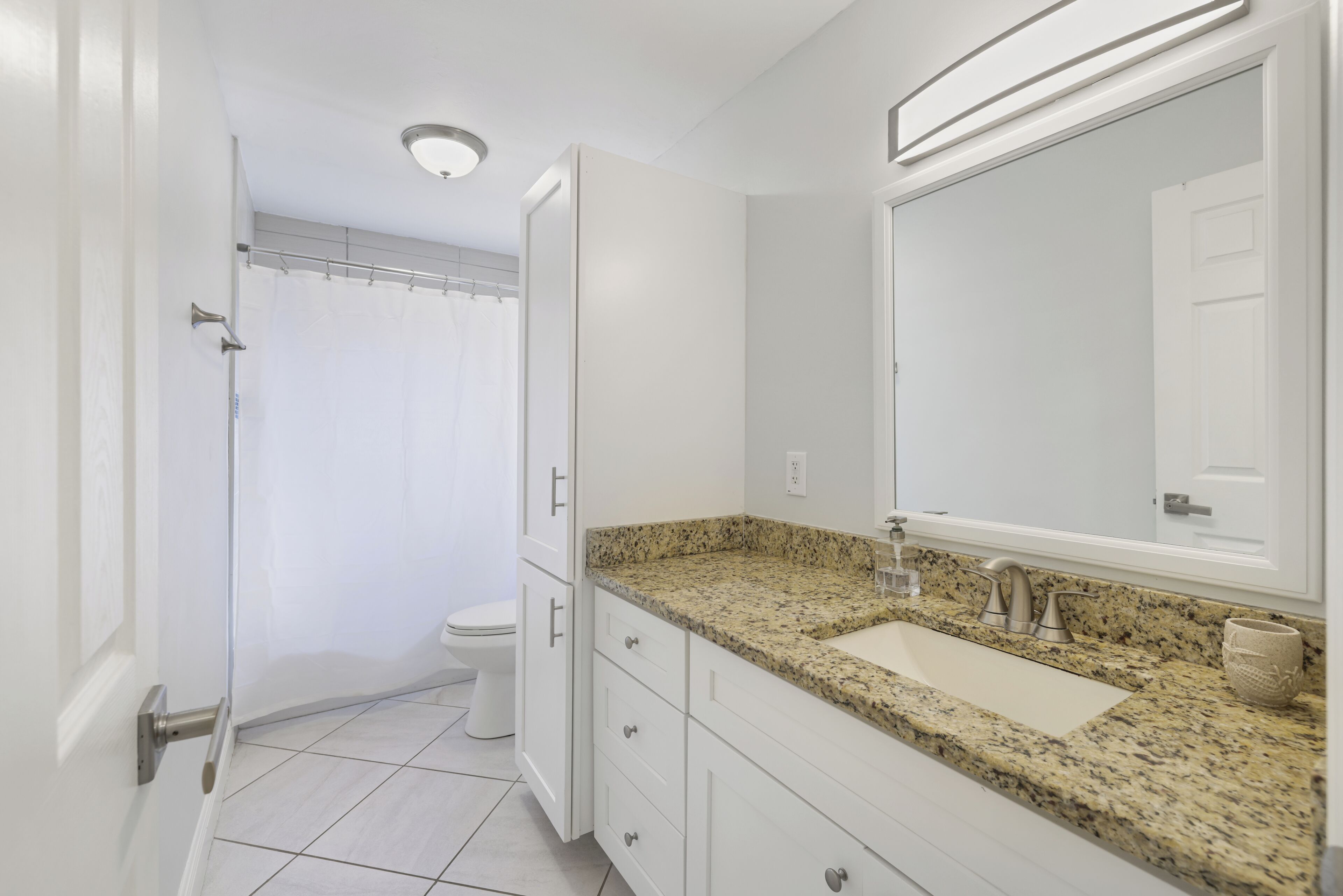 Une salle de bain avec des toilettes blanches, une douche dotée d’une porte en verre, un lavabo sur un plan de travail en granit et un grand miroir.