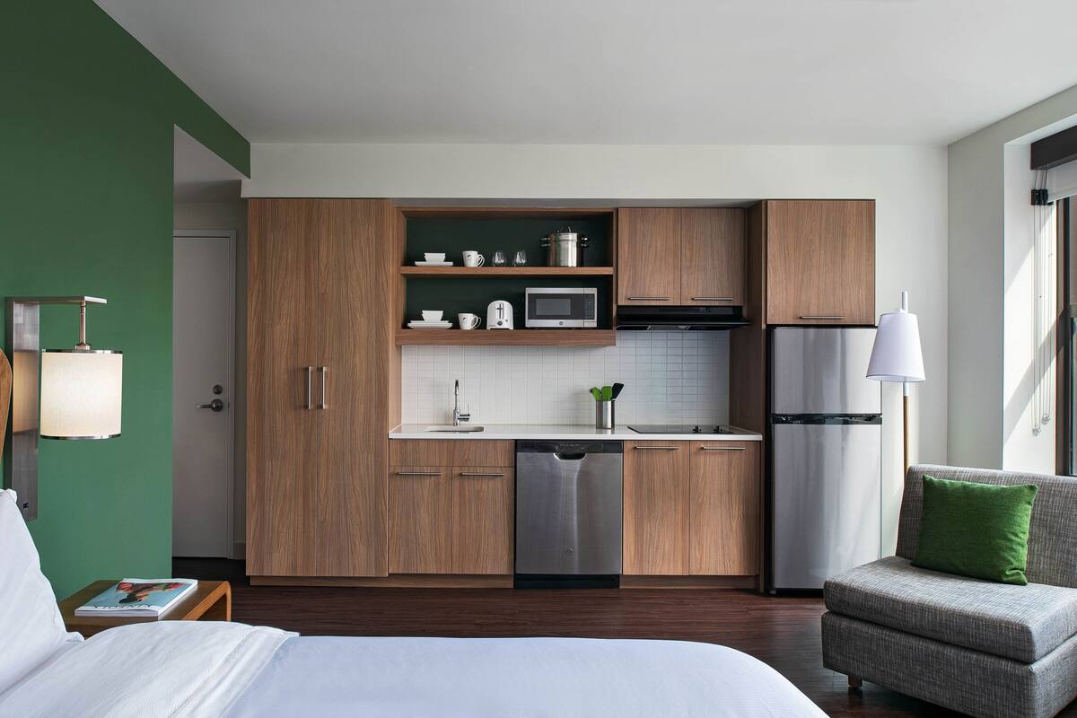 A modern hotel room with a kitchenette, a bed, a small armchair, and a bedside table with a book.