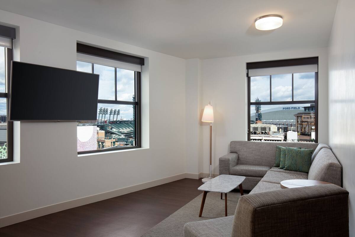A modern living room with a large flat-screen TV mounted on the wall, a grey sofa with green cushions, a small white coffee table, and a floor lamp.