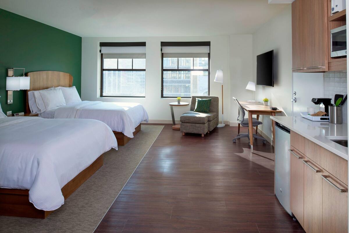 A hotel room with two beds, a small kitchenette, a dining area, and a view of the city.