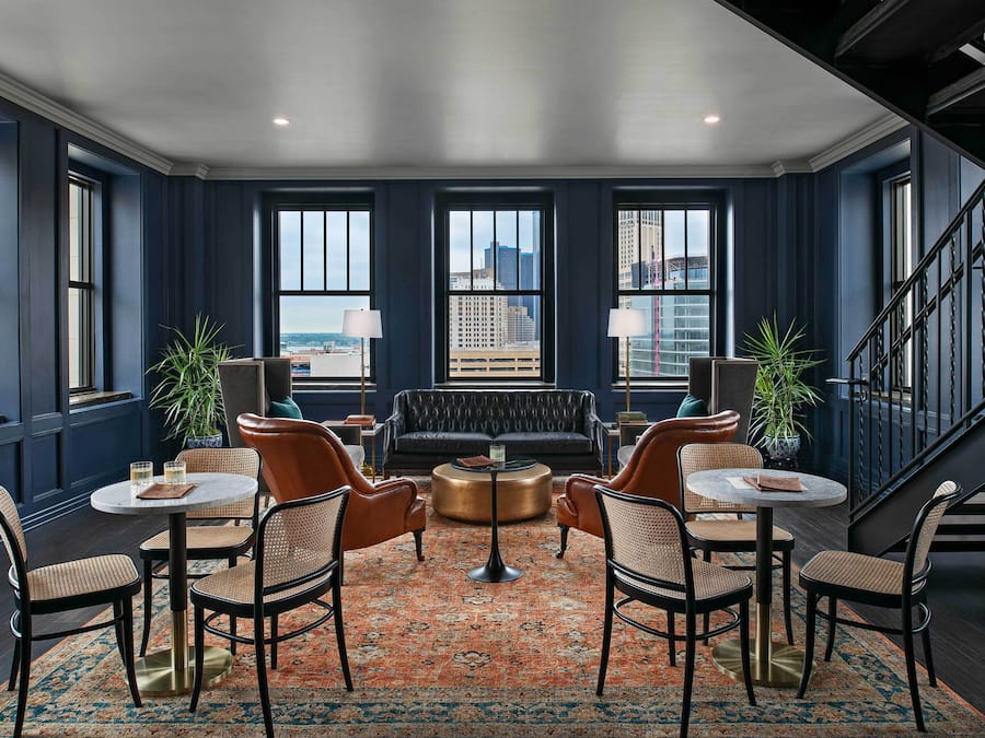 A spacious lounge area with a dark blue wall, large windows, a leather sofa, round tables, and chairs.