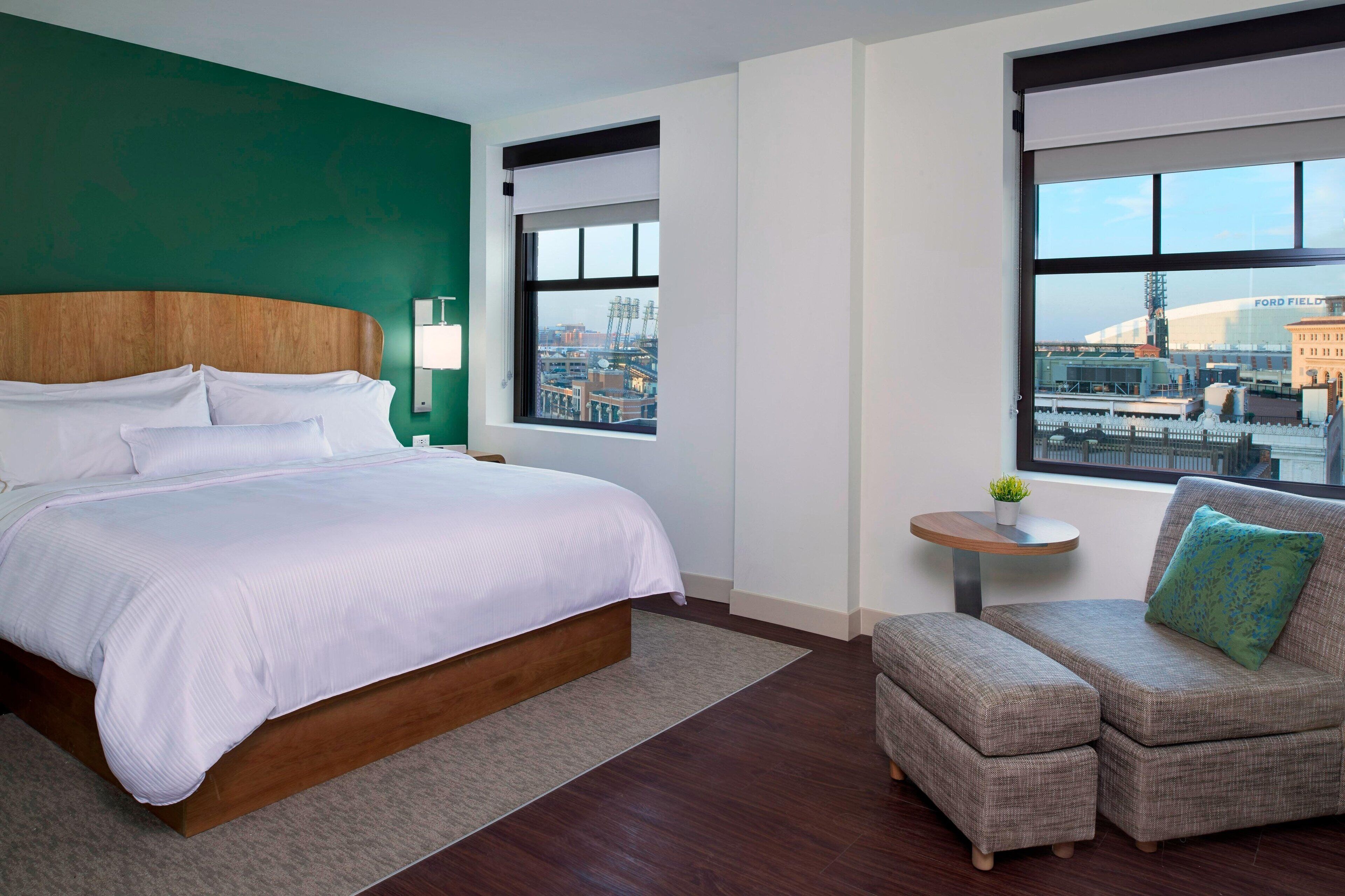 A hotel room with a large bed, a small sofa, a round table with a plant, and a view of a stadium through the windows.