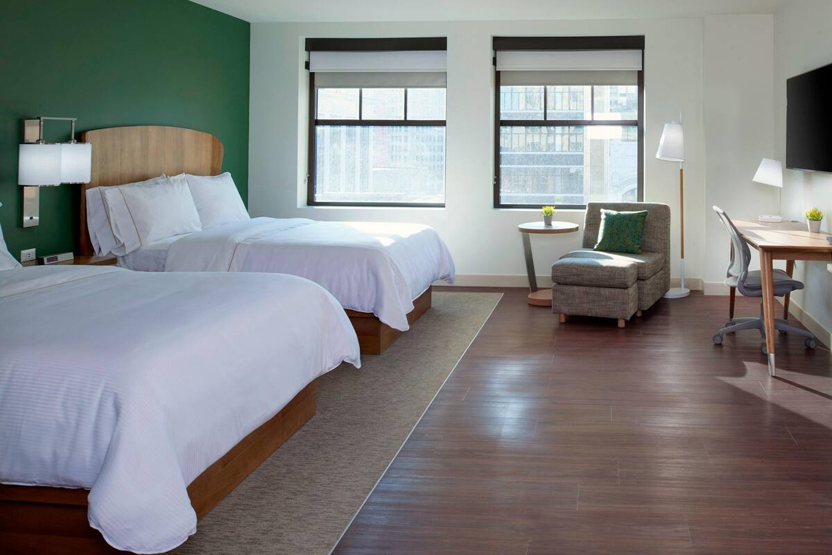 A hotel room with two beds, a desk, a chair, a TV, and two windows.