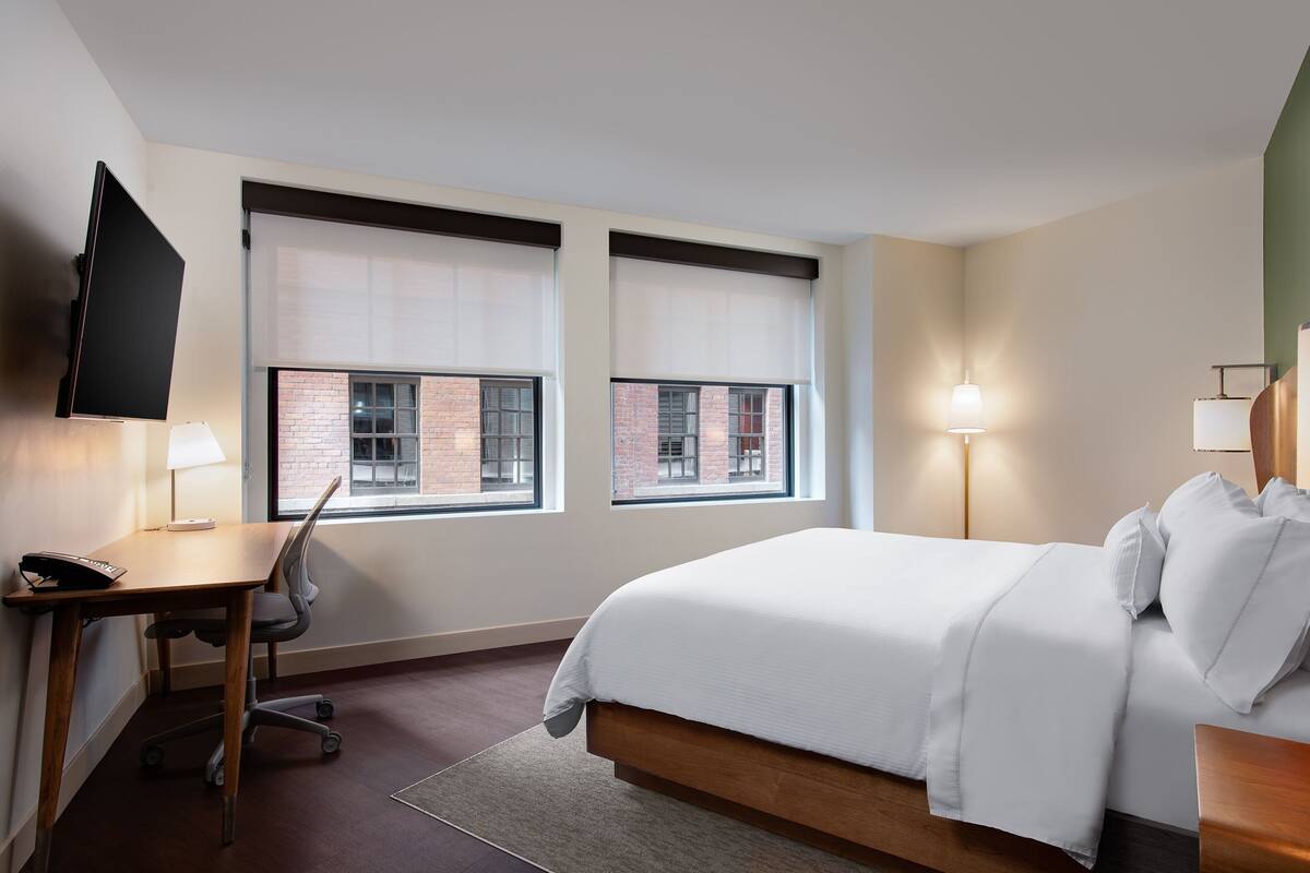 A hotel room with a bed, desk, chair, and two windows with blinds.