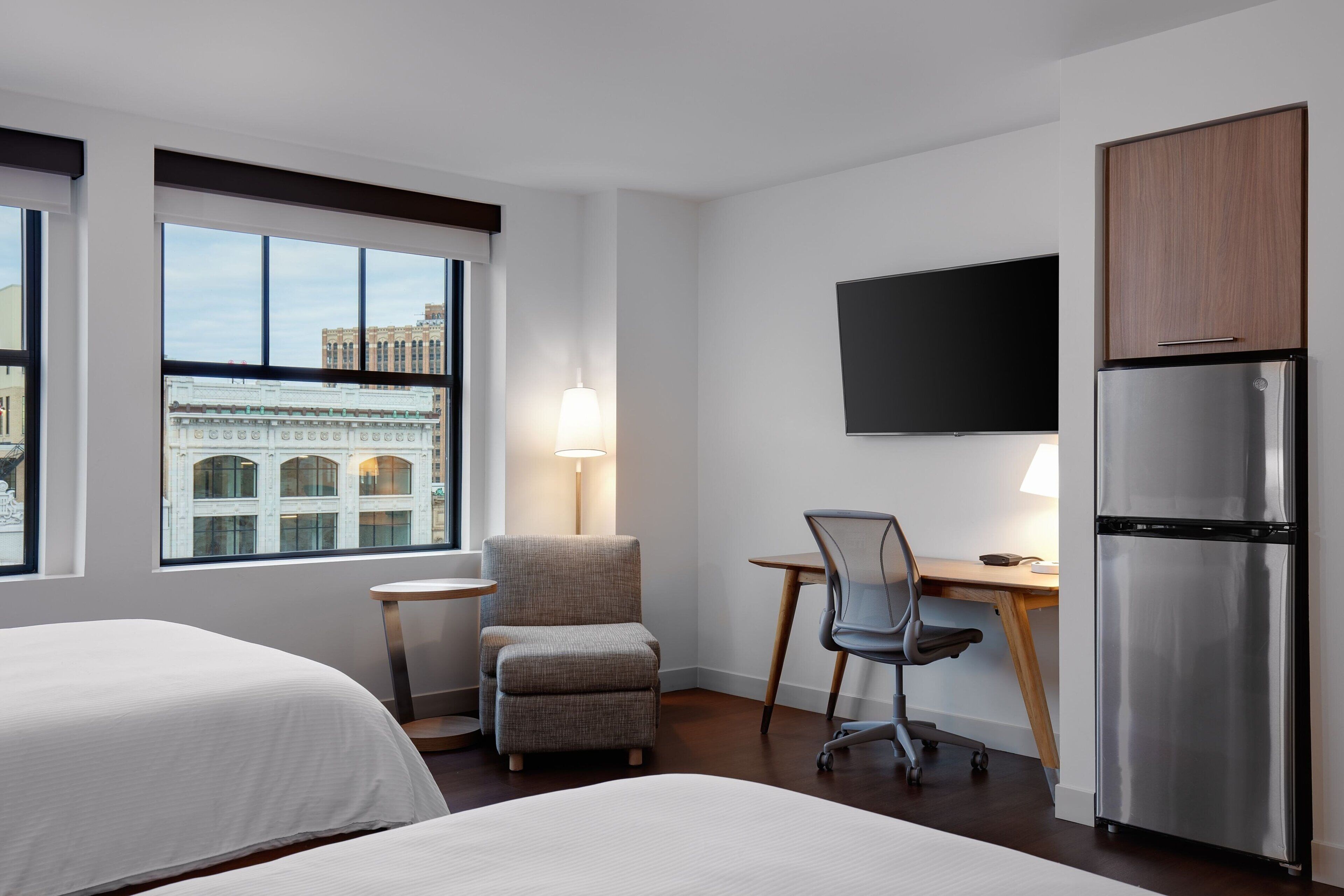 A hotel room with two beds, a desk with a chair, a refrigerator, a TV mounted on the wall, and a window with a view of buildings.
