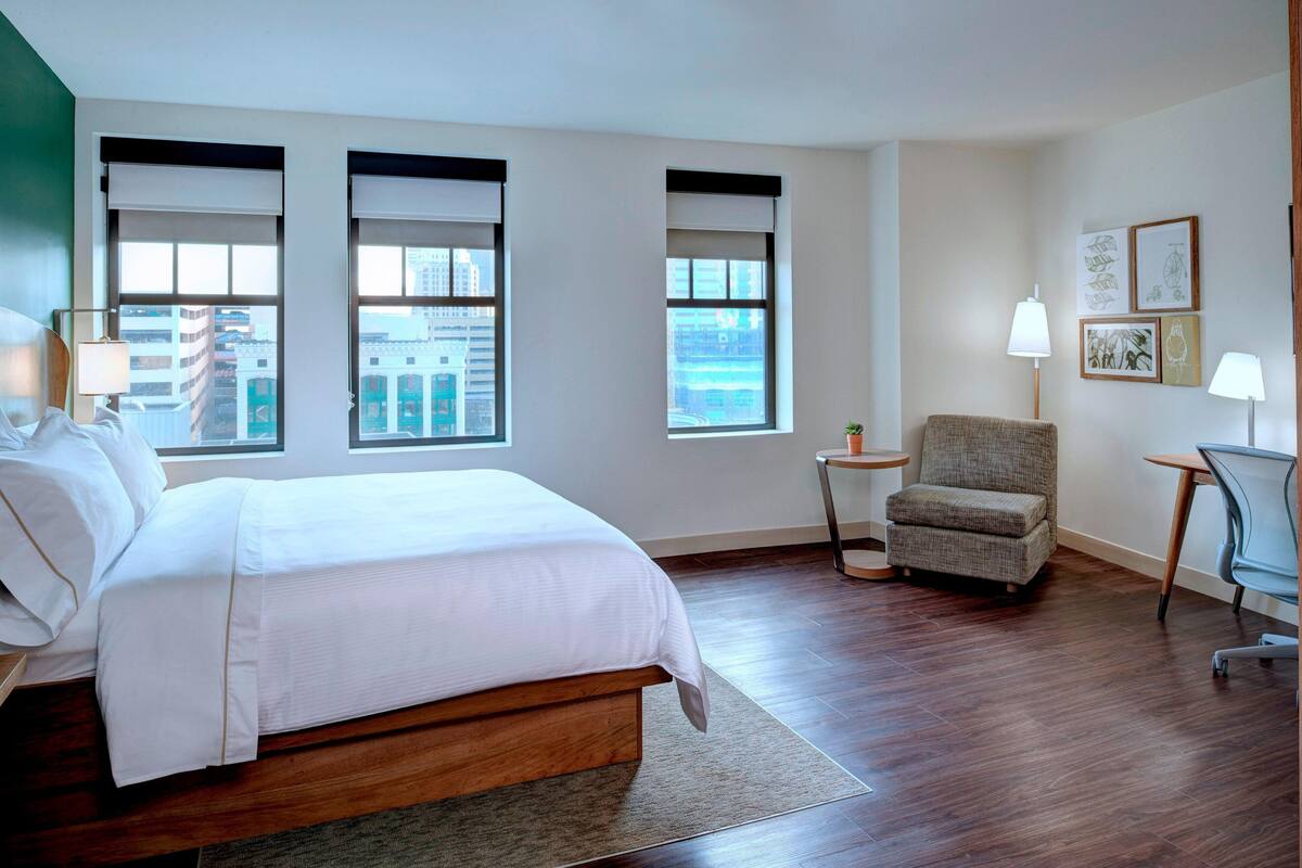 A hotel room with a large bed, two windows with blinds, a chair, a small table, and a lamp.