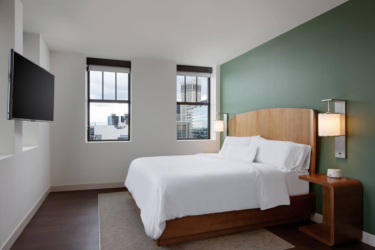 A modern hotel room with a large bed, two windows with city views, a flat-screen TV, and a nightstand with a lamp.
