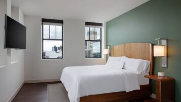 Une chambre d’hôtel moderne dotée d’un grand lit, de deux fenêtres donnant sur la ville, d’un téléviseur à écran plat et d’une table de chevet avec une lampe.