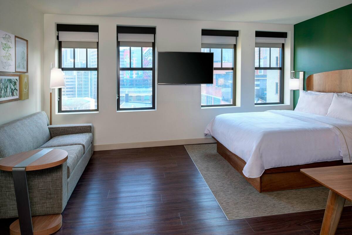 A hotel room with a bed, a sofa, a TV, and a window with a city view.