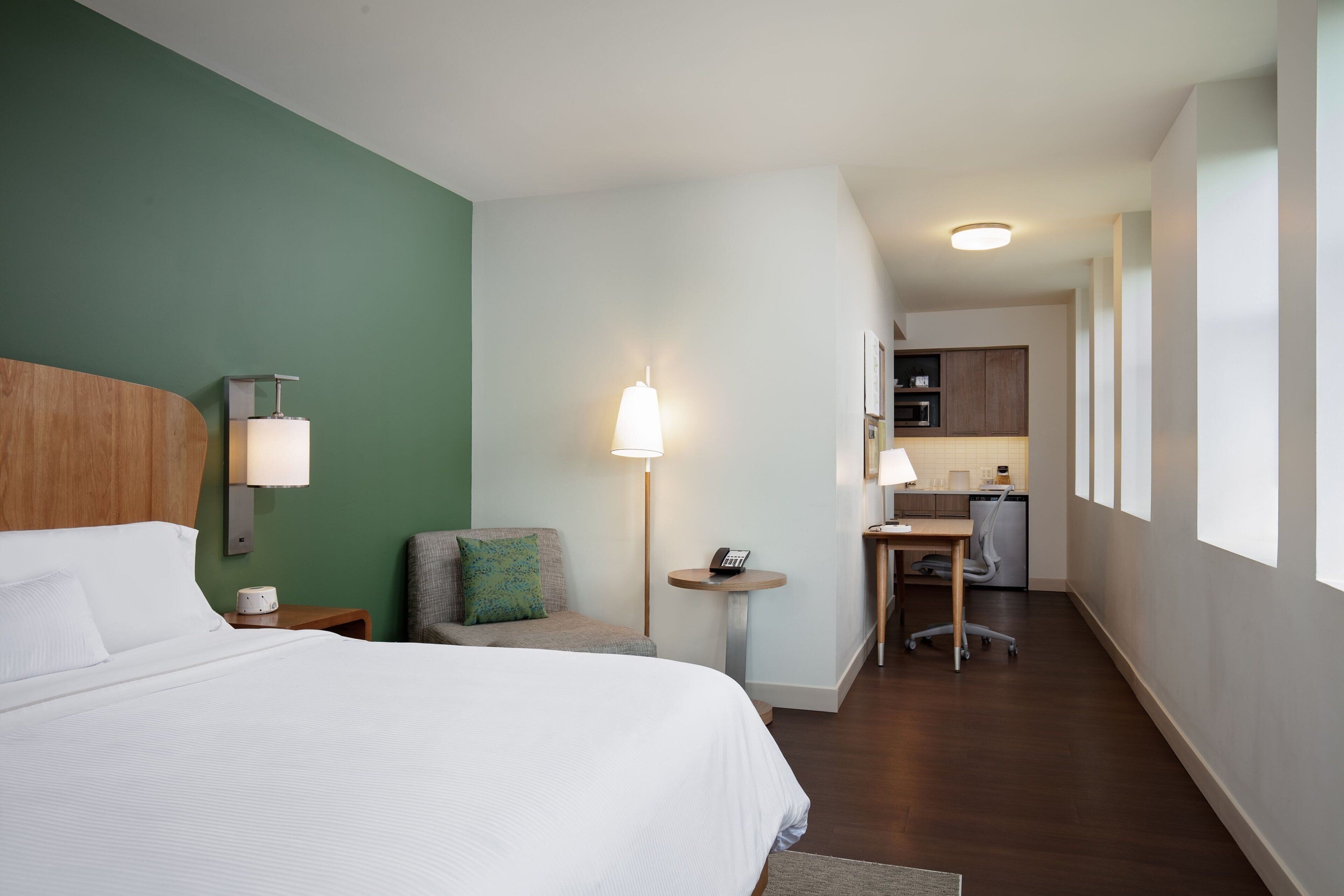 A hotel room with a bed, a desk, a chair, a lamp, and a view of the kitchen.