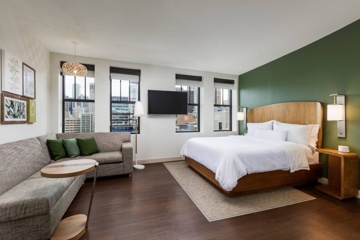A modern hotel room with a large bed, a sofa, a TV, and a city view.