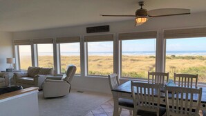 A spacious living room with a view of the beach, a dining area with white chairs, and a ceiling fan. - Pacific Dreams Beach House- Beach front! Gorgeous views! Meticulously sanitized! (Grayland)