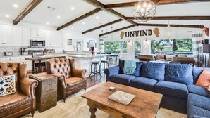 A spacious living area with a sofa, coffee table, and leather armchairs. A kitchen with white cabinets and a microwave is visible in the background.