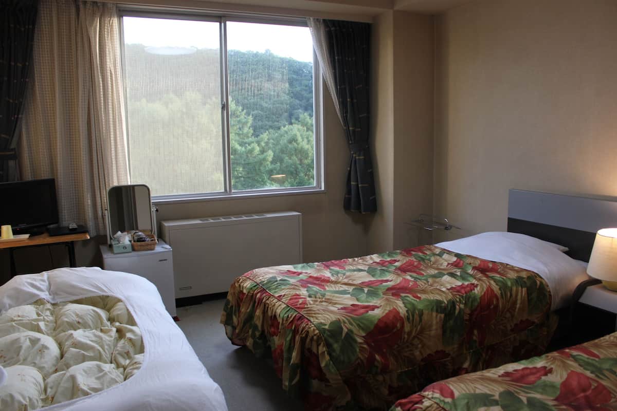 Quarto de hotel com duas camas, uma janela com cortinas, uma escrivaninha com abajur e uma televisão.