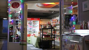 A brightly lit store with shelves stocked with various products, a display counter, and a refrigerated section.
